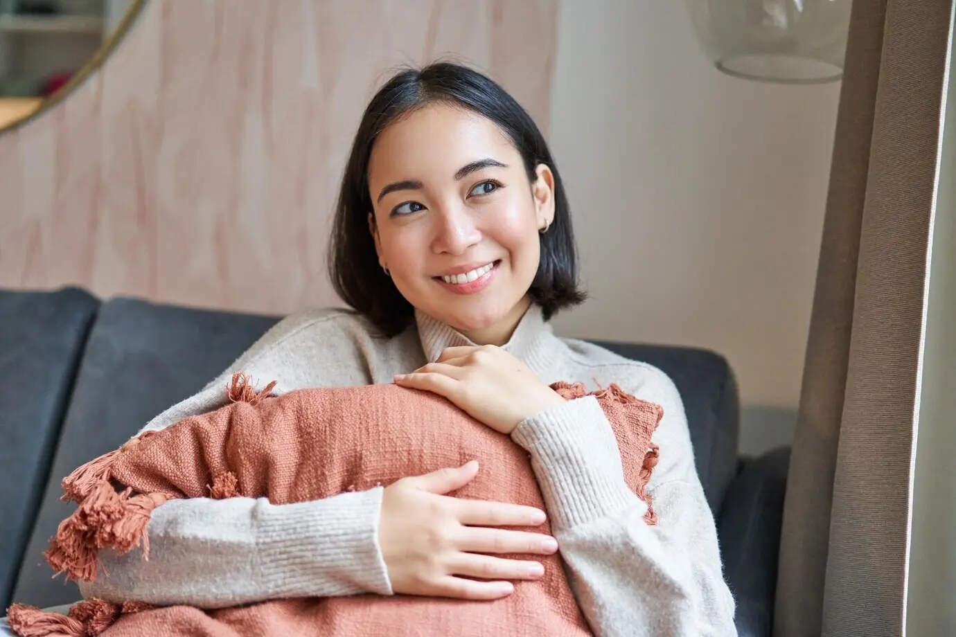 Konzept Frauen und Wohlbefinden: Eine junge Frau in warmer Kleidung sitzt zu Hause auf dem Sofa, umarmt ein Kissen und lächelt.